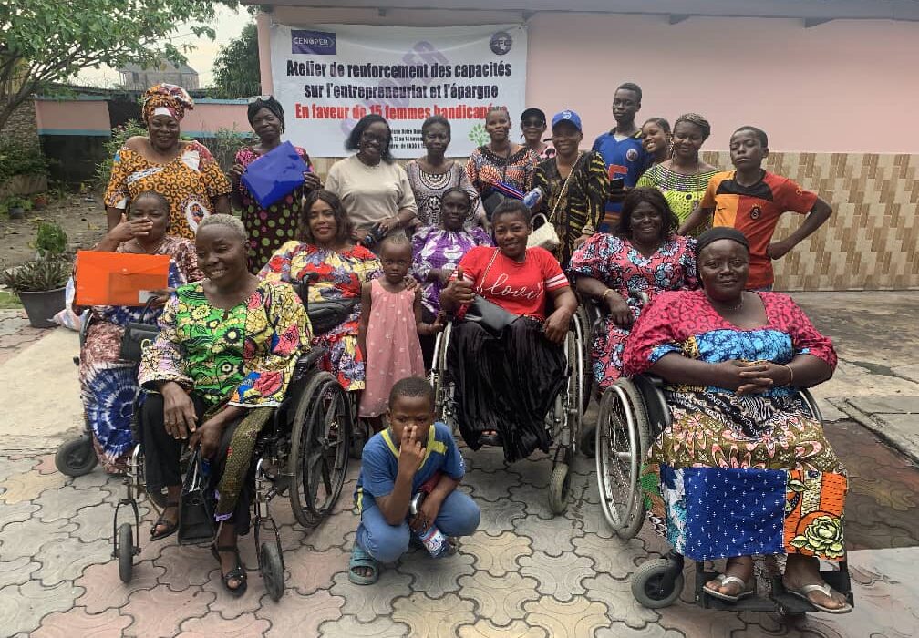 A diverse group of women, some in wheelchairs, poses together outside a building with a banner about empowerment for women with disabilities. They are smiling, creating a positive and supportive atmosphere.