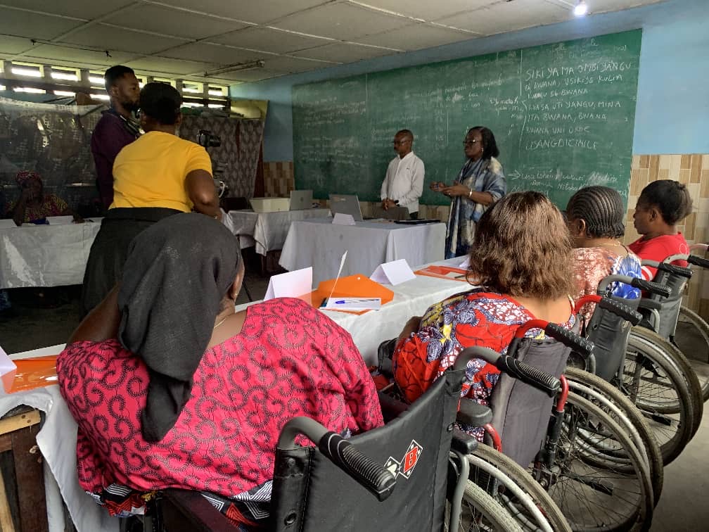 A classroom with people in wheelchairs facing a chalkboard. Two presenters stand at the front, engaging with the group, creating an inclusive learning environment.