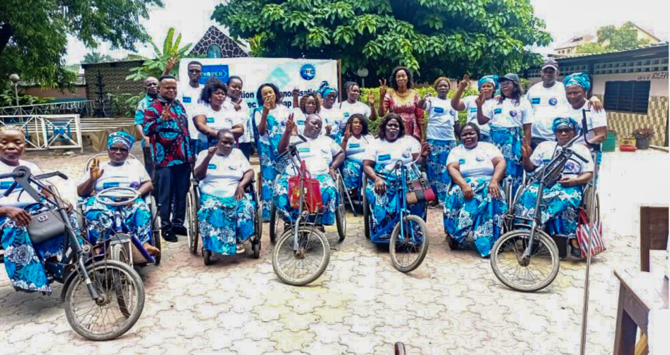Groupe de femmes africaines handicapées posant ensemble en extérieur lors d’un événement communautaire, certaines en fauteuil roulant et d’autres utilisant des tricycles adaptés, portant des tenues traditionnelles assorties bleu et blanc avec foulards, souriant et faisant des signes de paix devant une bannière d’organisation, dans une cour arborée avec bâtiments en arrière-plan, illustrant inclusion, autonomisation des femmes et mobilité assistée en Afrique.