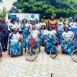 Groupe de femmes africaines handicapées posant ensemble en extérieur lors d’un événement communautaire, certaines en fauteuil roulant et d’autres utilisant des tricycles adaptés, portant des tenues traditionnelles assorties bleu et blanc avec foulards, souriant et faisant des signes de paix devant une bannière d’organisation, dans une cour arborée avec bâtiments en arrière-plan, illustrant inclusion, autonomisation des femmes et mobilité assistée en Afrique.