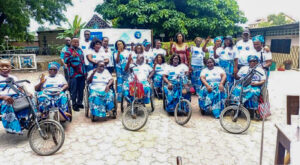 Groupe de femmes africaines handicapées posant ensemble en extérieur lors d’un événement communautaire, certaines en fauteuil roulant et d’autres utilisant des tricycles adaptés, portant des tenues traditionnelles assorties bleu et blanc avec foulards, souriant et faisant des signes de paix devant une bannière d’organisation, dans une cour arborée avec bâtiments en arrière-plan, illustrant inclusion, autonomisation des femmes et mobilité assistée en Afrique.